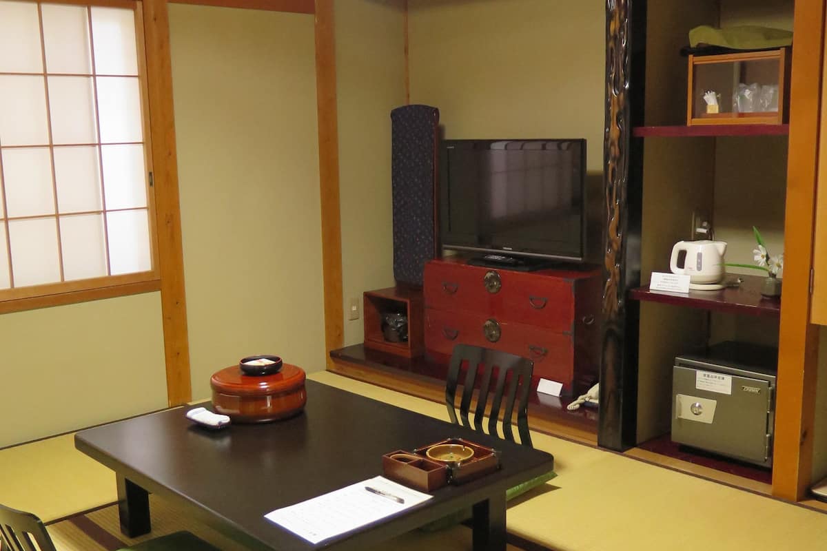 A traditional Japanese-style room with tatami flooring, a low table with a cushion, a television, a cabinet, and a small safe.