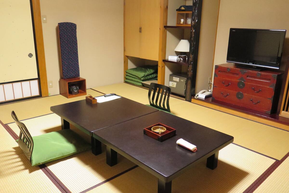 A tatami mat room with a low table, chairs, a television, and a red chest of drawers.