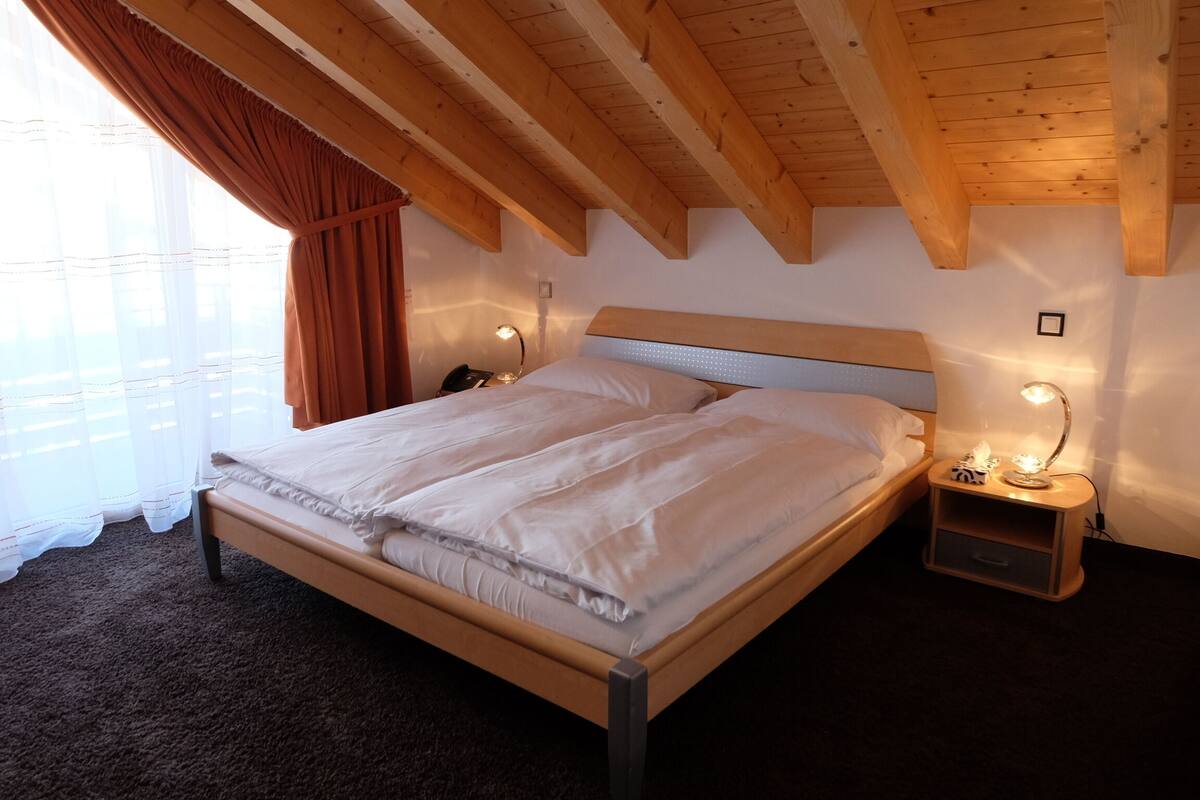 A bedroom with a wooden ceiling, a bed with white bedding, a nightstand with a lamp, and a window with curtains.