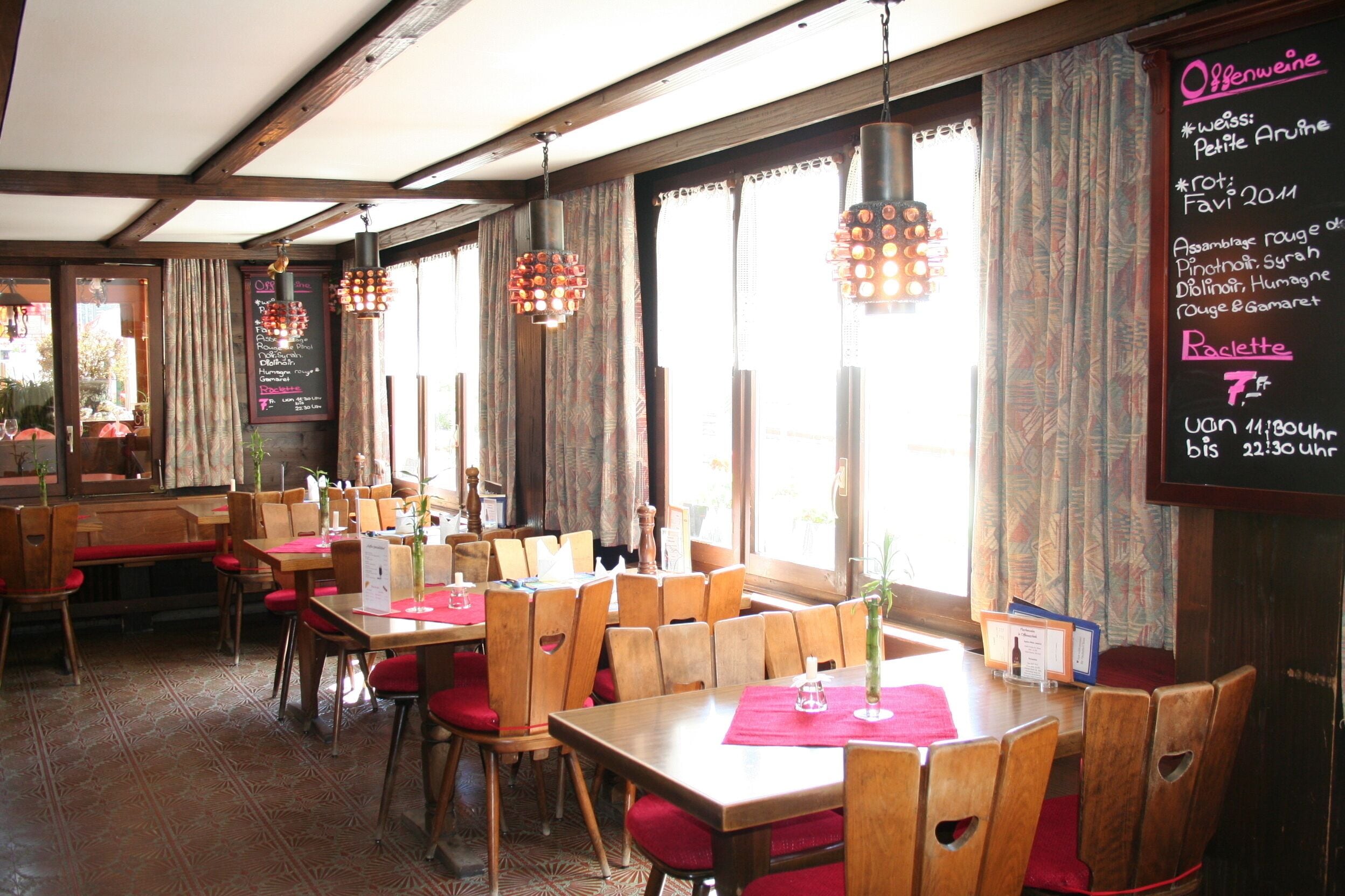 Ein Restaurantinneres mit Holztischen und -stühlen, einer Kreidetafel mit der Speisekarte und hängenden Pendelleuchten.