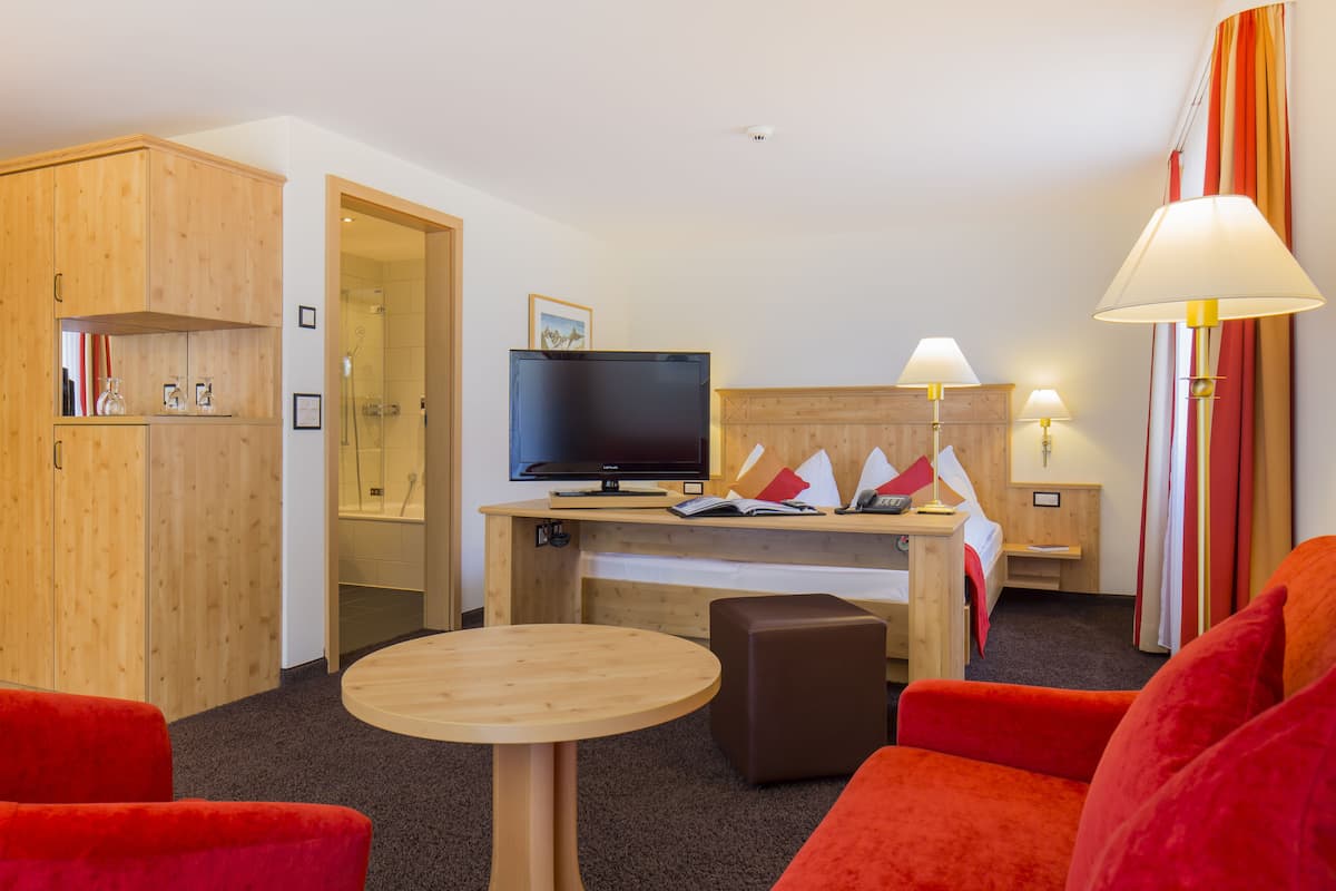 A hotel room with a wooden wardrobe, a television, a desk with a computer, a small round table, a sofa, and a lamp.