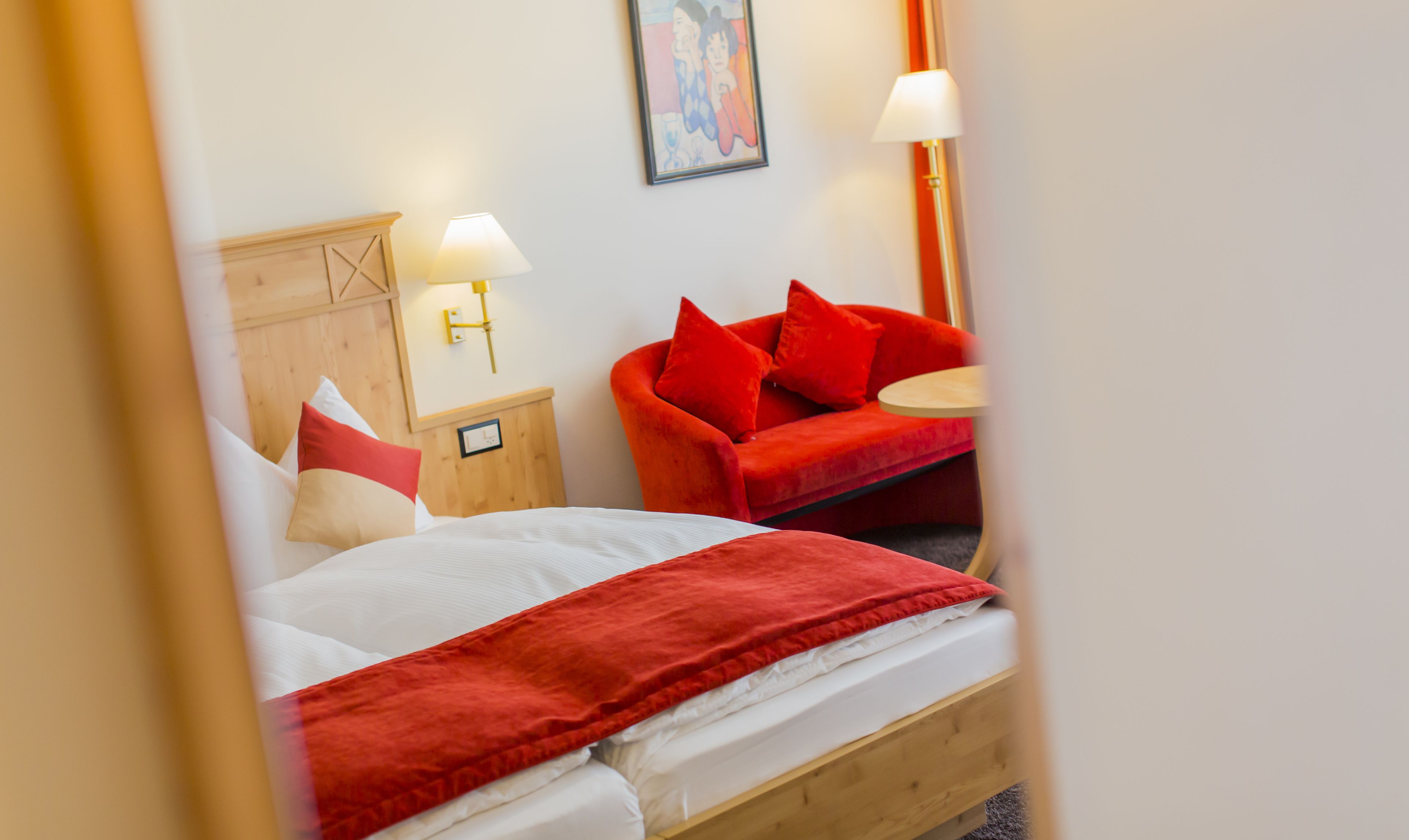 Quarto de hotel com uma cama de madeira, um sofá vermelho, uma mesa redonda pequena e uma luminária fixada na parede.
