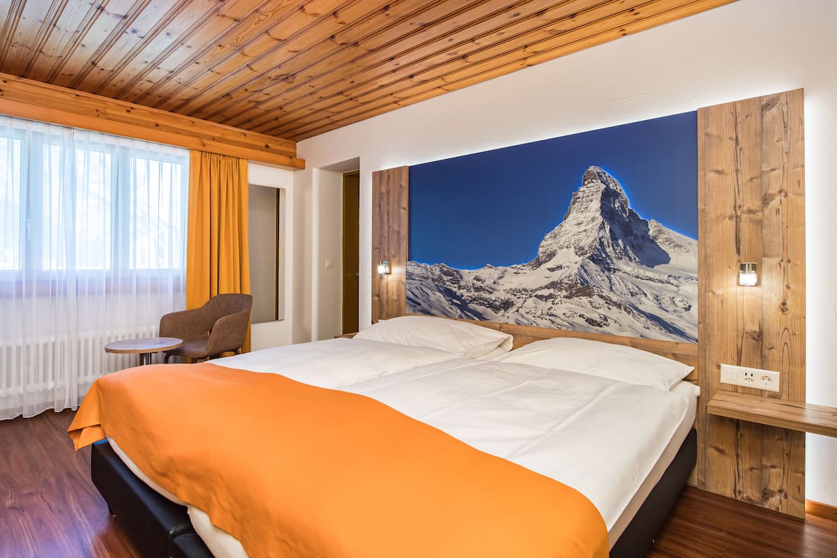A hotel room with a large bed, a wooden ceiling, a mountain painting on the wall, and a small table with a chair.