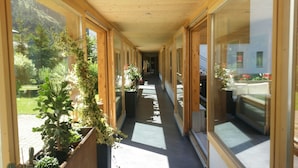Een gang met houten wanden en grote ramen, die uitkomt in een kamer met een bed en uitzicht op een tuin.