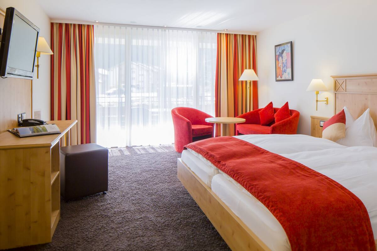 A hotel room with a bed, a desk, a chair, a small table, and a television.