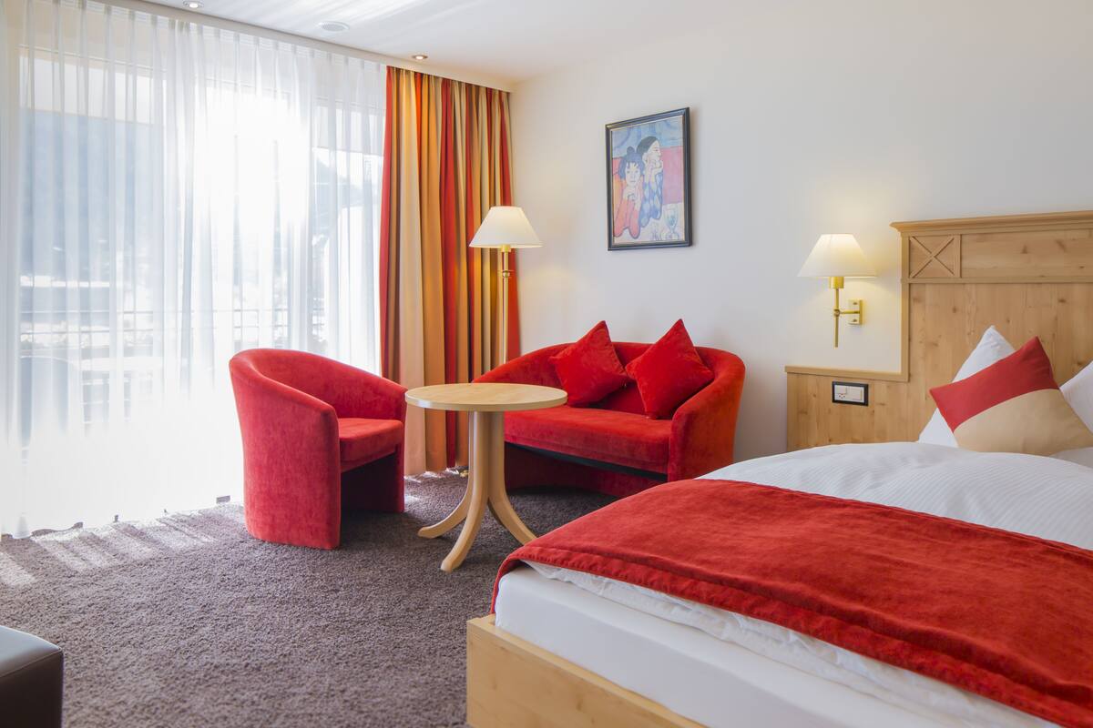 A hotel room with a bed, a red sofa, a red armchair, a small round table, and two wall-mounted lamps.