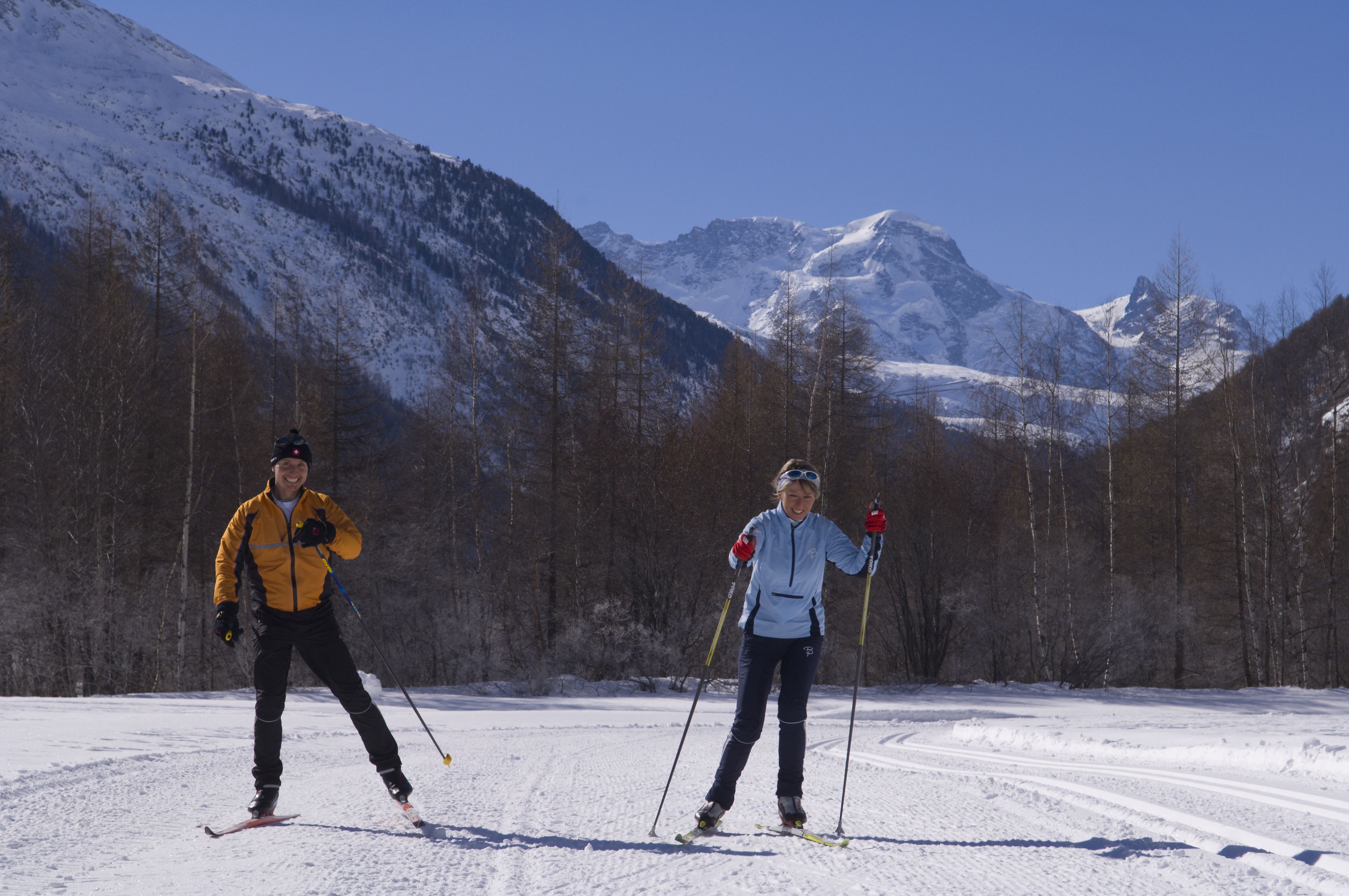 Two skiers on a snowy trail with mountains in the background.