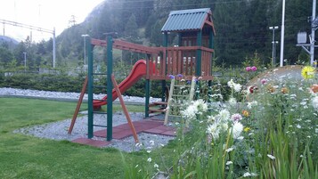 Un parque infantil con un tobogán rojo y una estructura de madera para escalar, rodeado de un jardín con diversas flores y un camino de grava.
