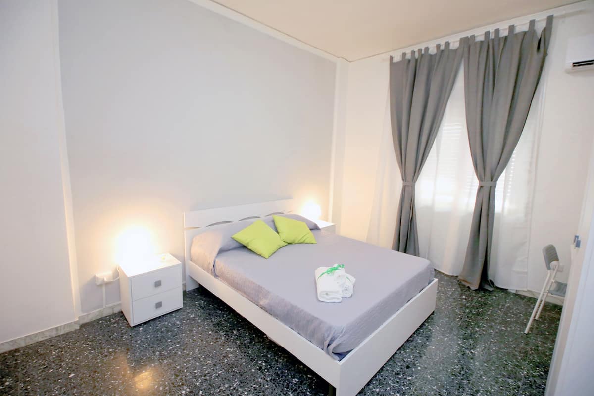 A neatly made bed with a grey bedspread and two pillows, a white bedside table with a lamp, and a window with grey curtains.