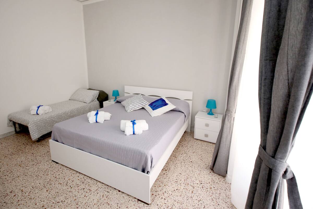 A hotel room with a large bed, a smaller bed, two bedside tables, and a window with curtains.