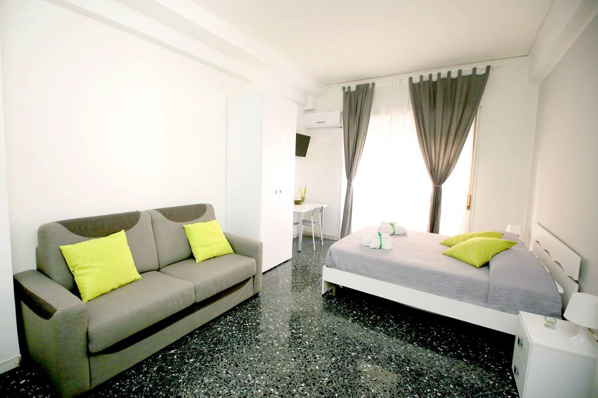 A modern hotel room with a grey sofa, a bed with grey bedding and green pillows, a small white bedside table, and a window with curtains.
