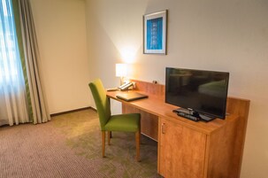 Una camera d'albergo con una scrivania in legno, una sedia verde, una televisione, una lampada e un quadro appeso al muro.
