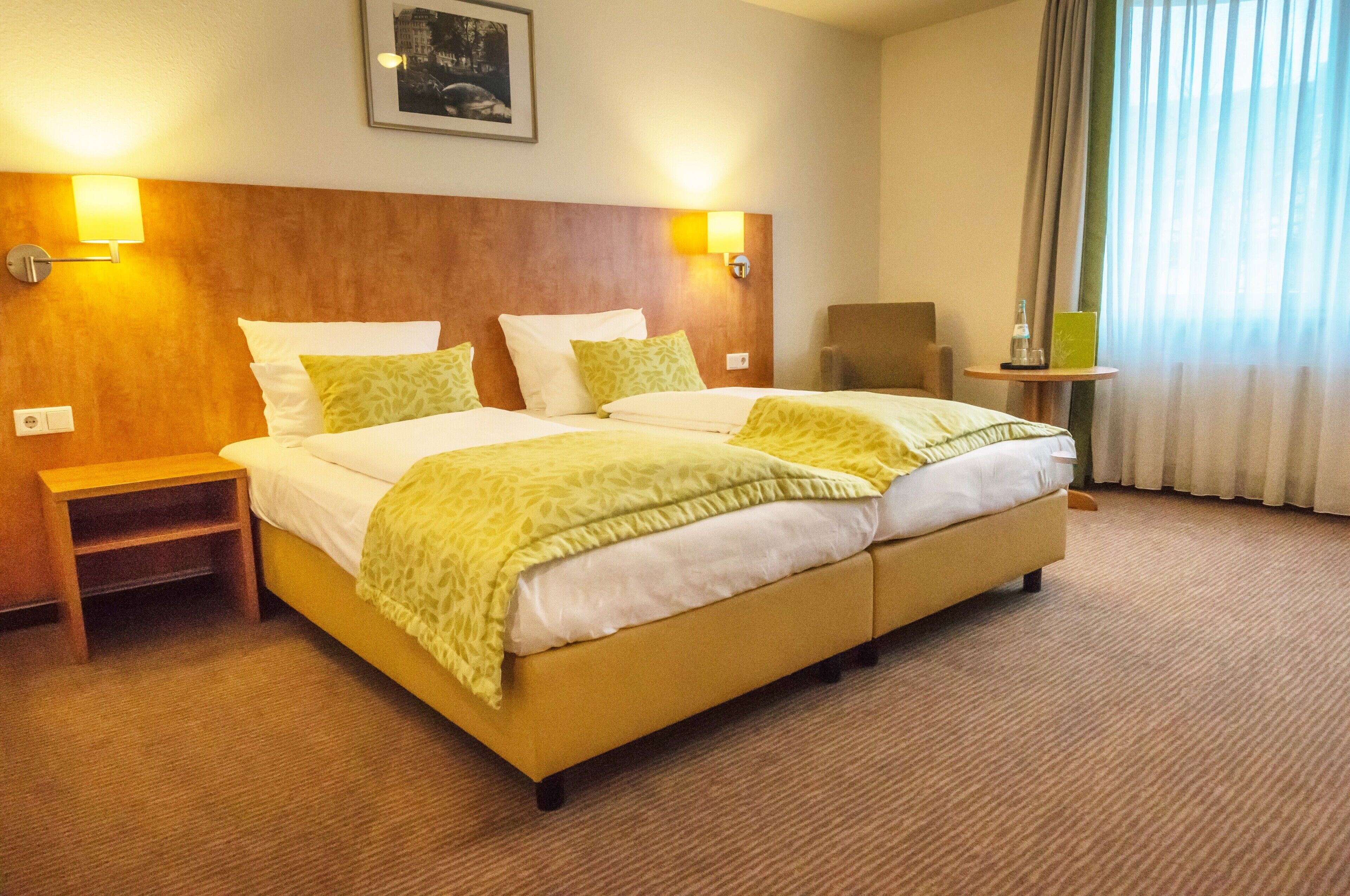 A hotel room with a double bed, two bedside tables, a chair, a small table, and a window with curtains.