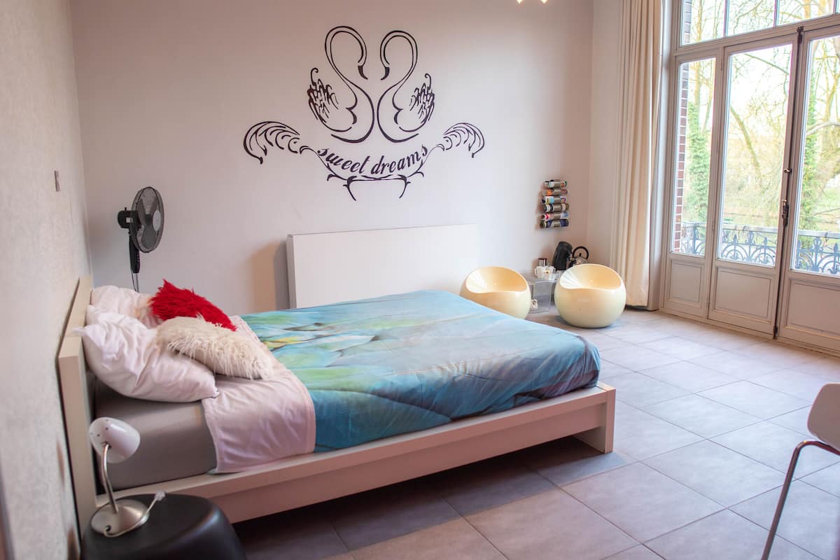 Quarto moderno com cama, ventilador fixado na parede, arte decorativa e janela com vista para árvores.