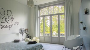A bedroom with a bed, a chair, a fan, and a window with curtains. - Vegan b&b AM/PM (Bruges)