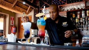 Een barman achter de bar, met een brandblusser en diverse drankflessen op de achtergrond.