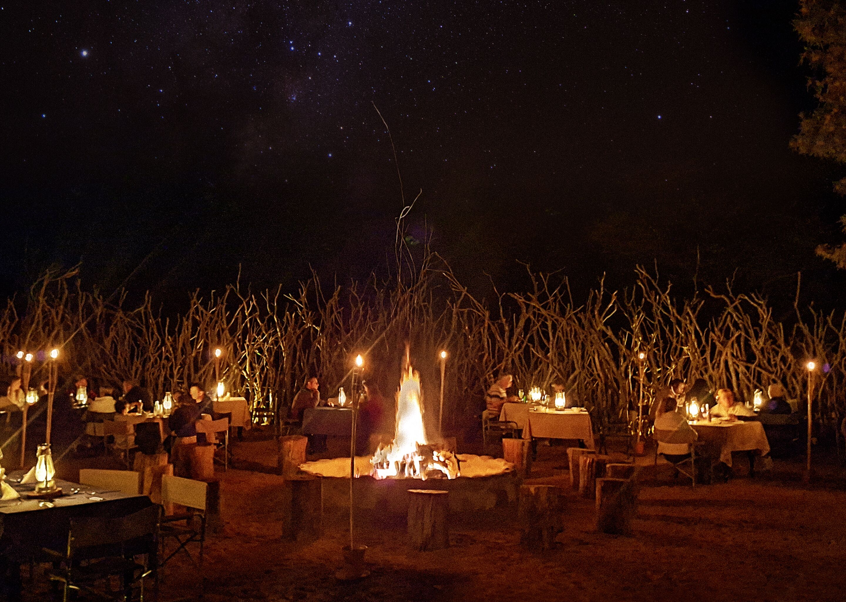 An outdoor dining area with tables arranged around a central fire pit, under a starry night sky.