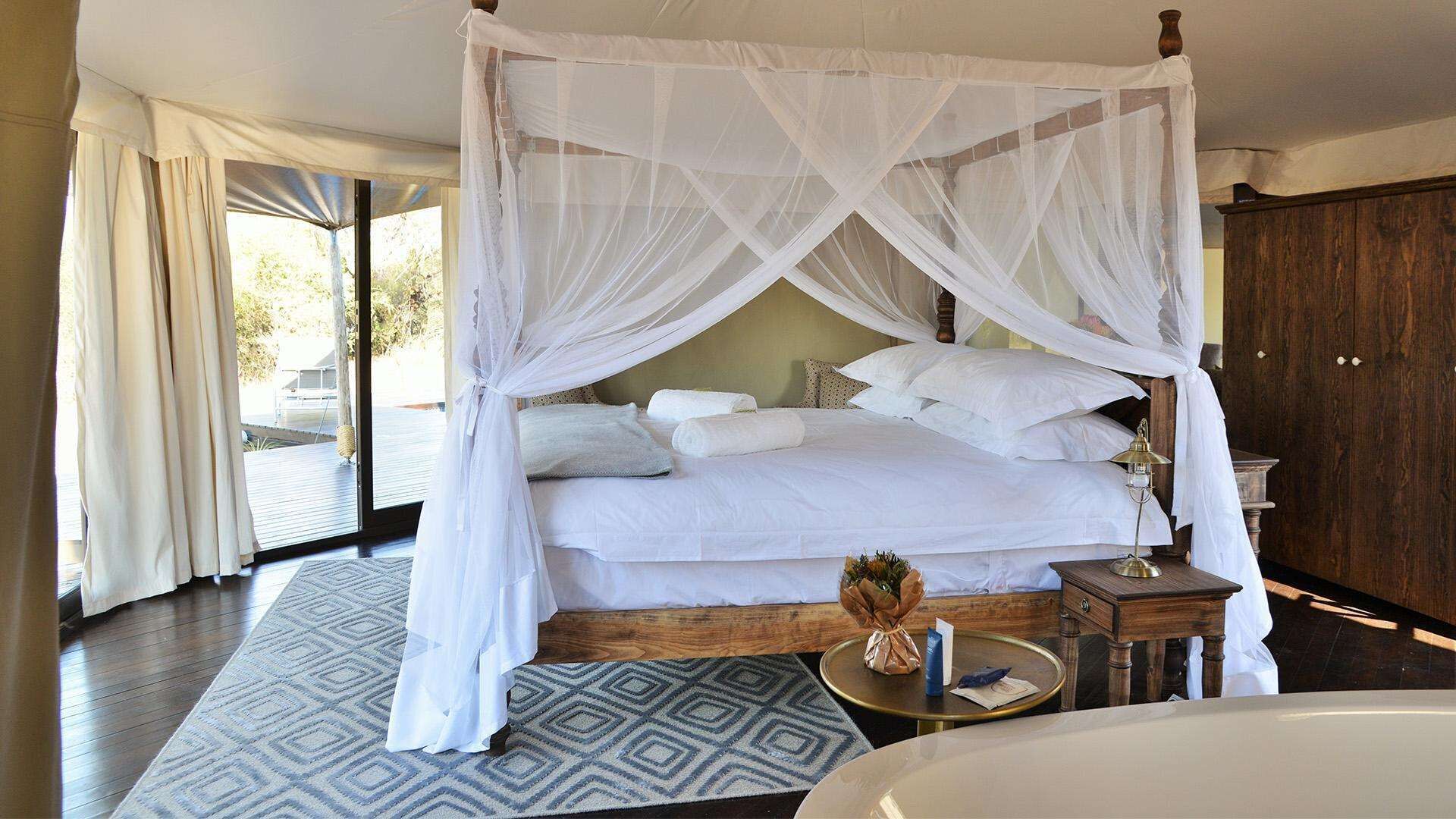 A four-poster bed with white linens, a bedside table with a lamp, and a view of the outdoors through large windows.