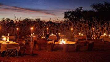 An outdoor dining area with tables and chairs arranged around a central fire pit, set against a backdrop of tall grasses and trees under a night sky.