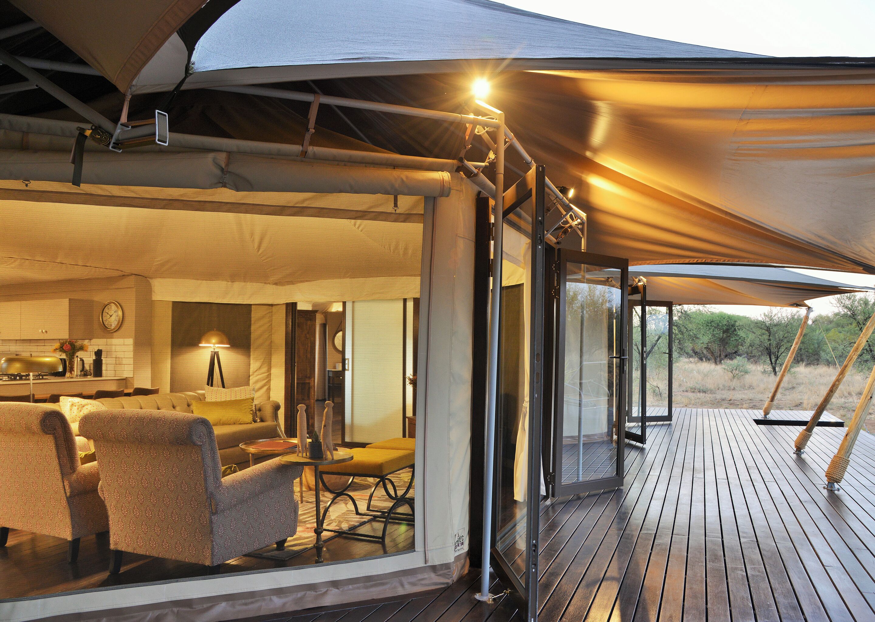 A spacious outdoor living area with a wooden deck, a dining set, and a kitchen area visible through a glass door.