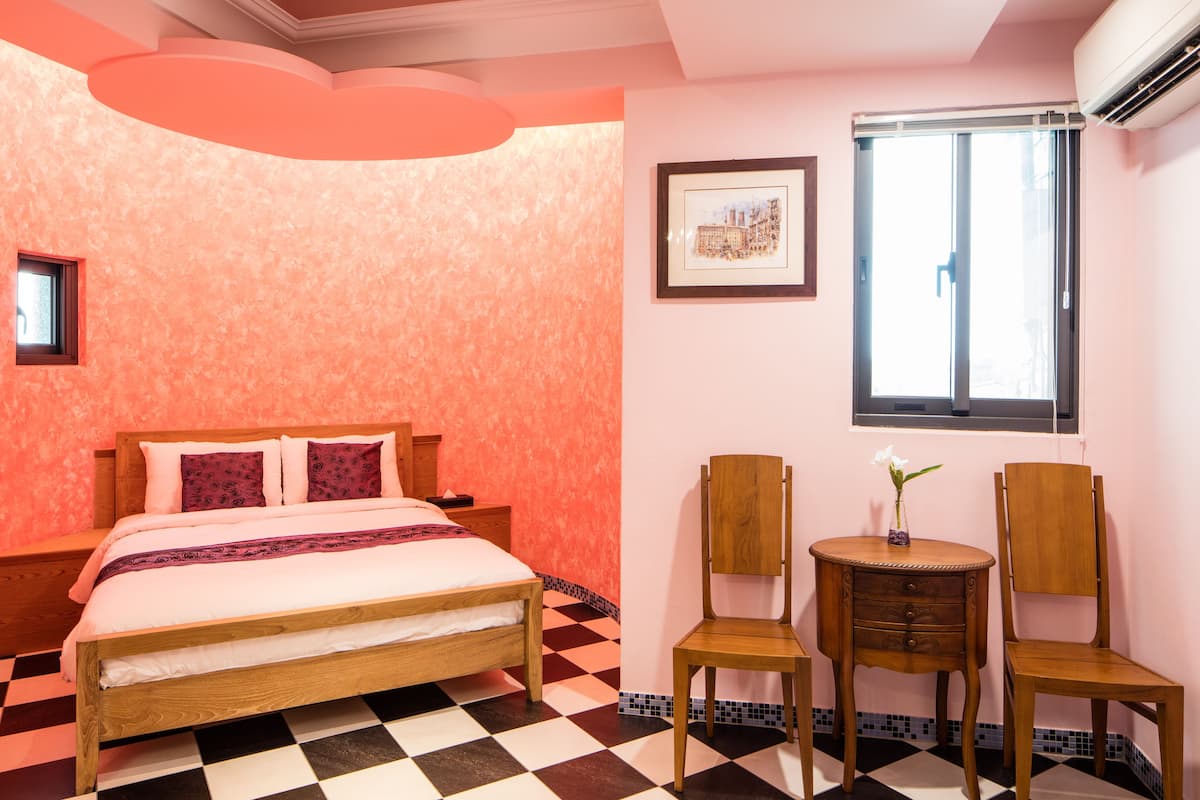 Een slaapkamer met een geruit vloerkleed, een bed met houten frame, twee houten stoelen, een kleine tafel met een vaas en een raam met jaloezieën.