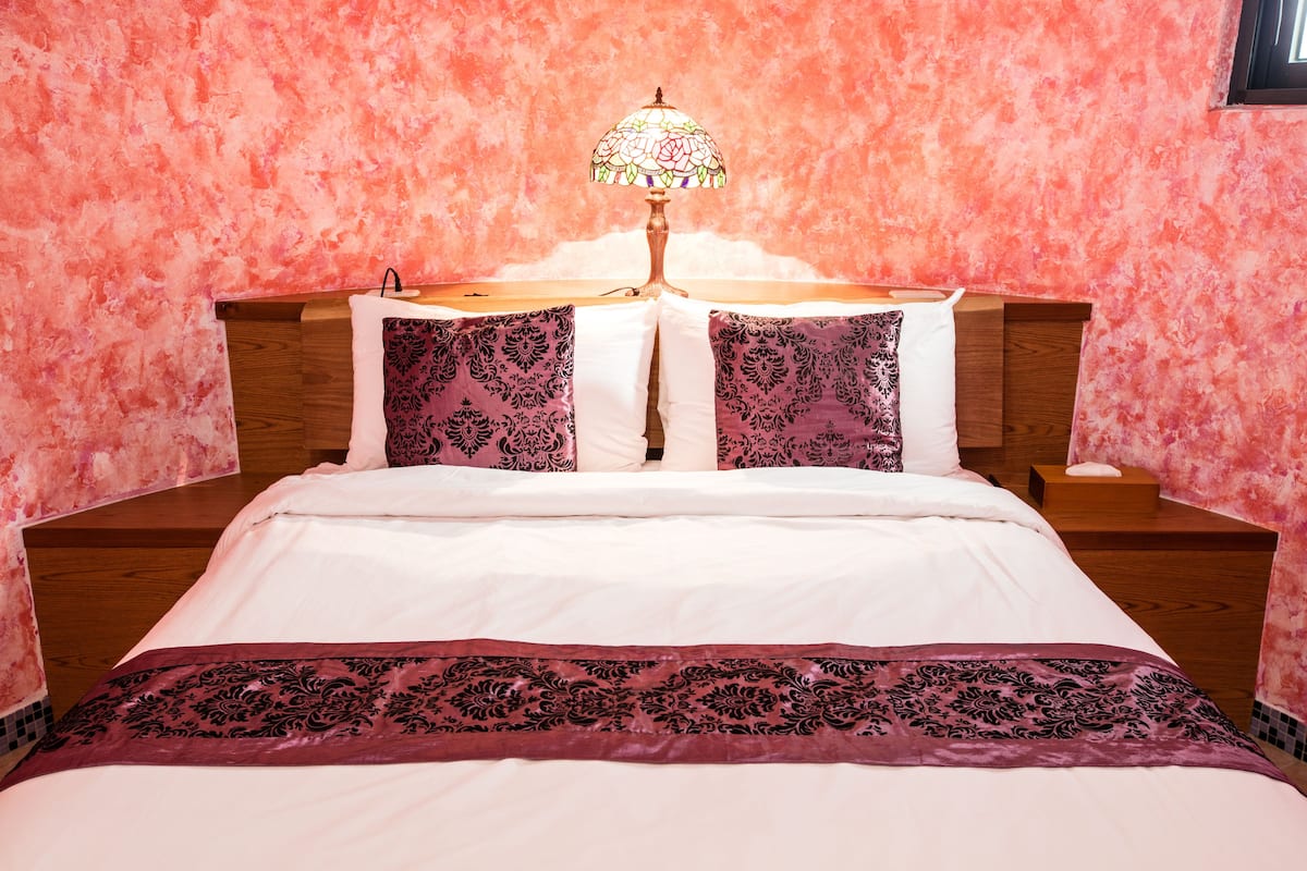 Een keurig opgemaakt bed met een wit en paars deken, twee bijpassende kussens, een nachtkastje met een lamp en een roze, gestructureerde muur.