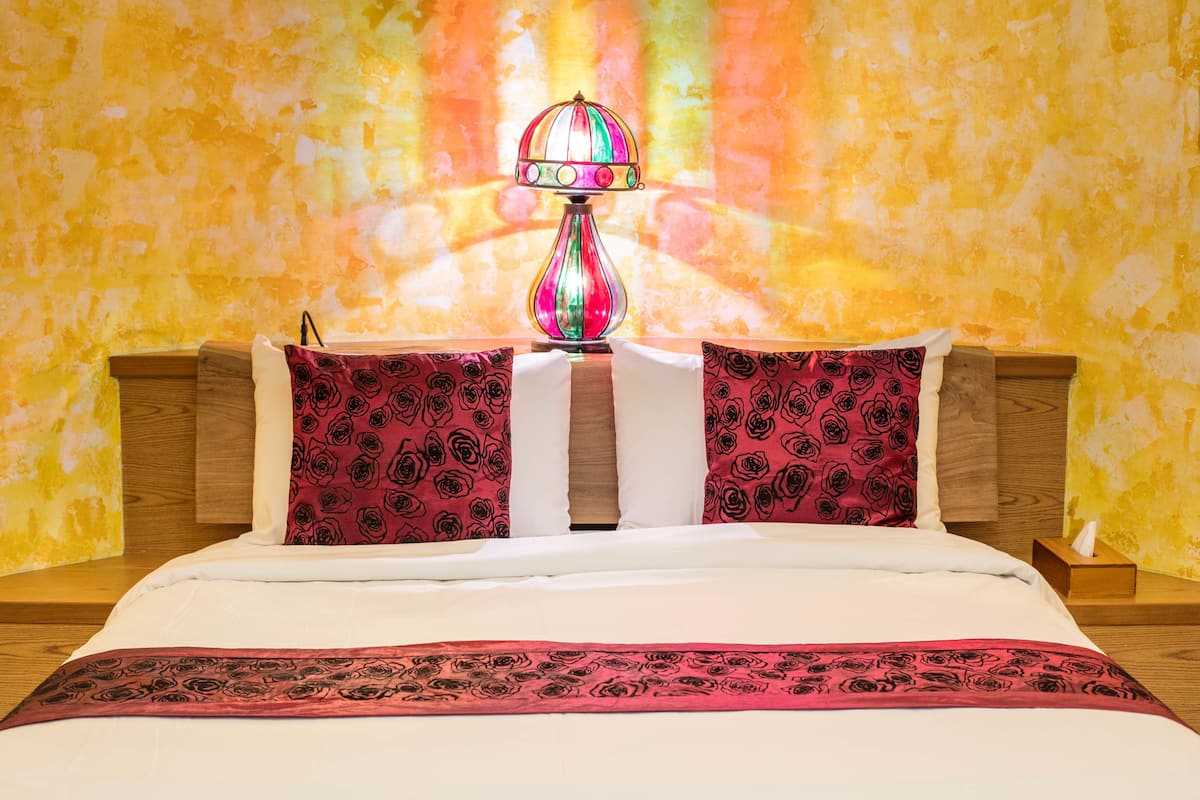 Een bed met een witte dekbedovertrek, twee rode kussens met een rozenprint en een rood-zwarte plaid. Een kleurrijke lamp staat op een houten nachtkastje naast het bed.