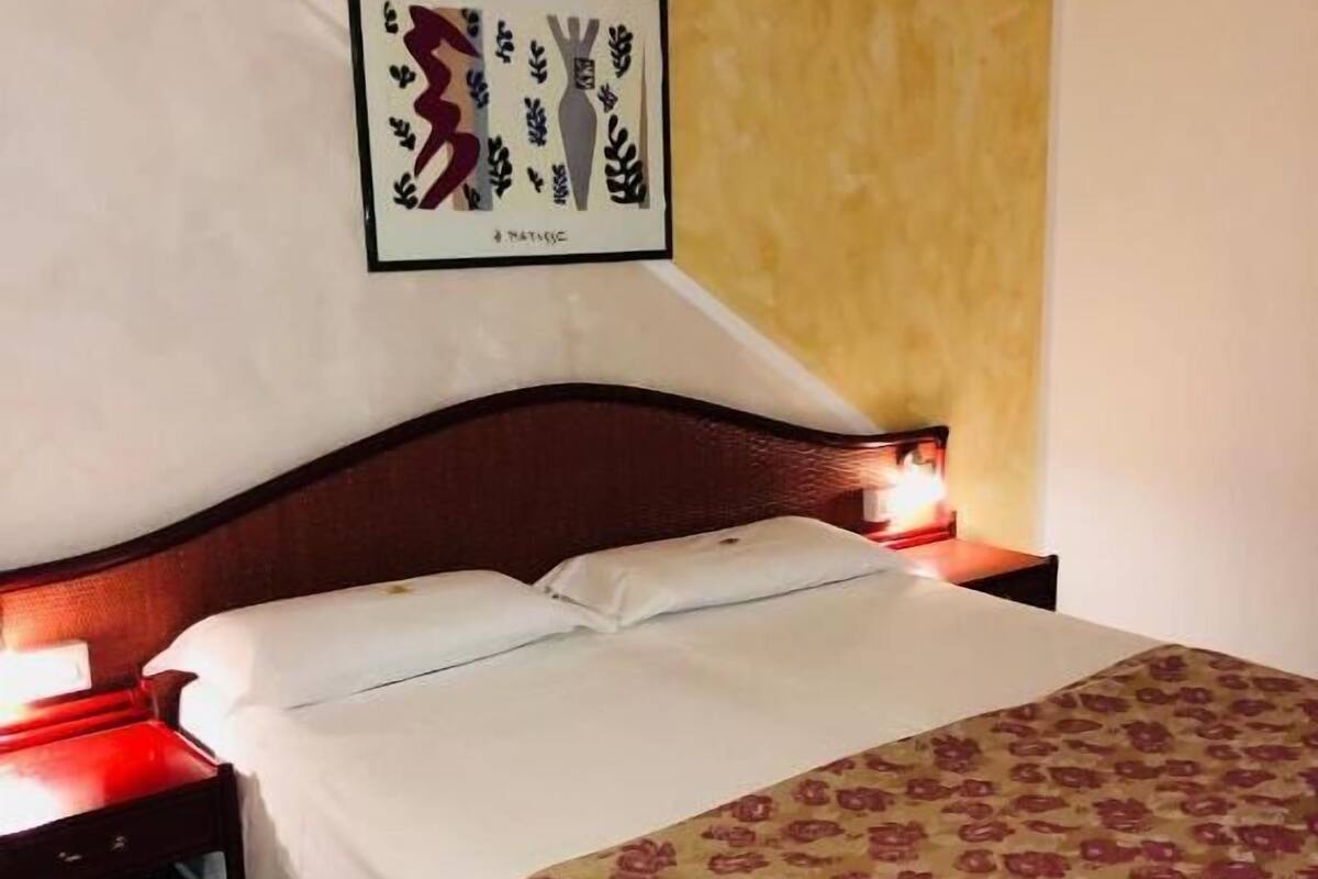 Un lit simple avec une couvre-lit à motifs floraux, une table de chevet avec une lampe rouge et une œuvre d’art encadrée accrochée au mur.