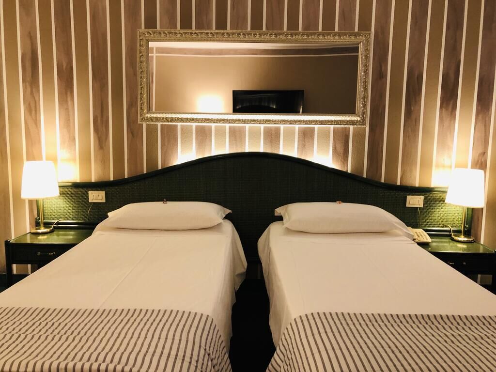 Camera d'albergo con due letti, un grande specchio sopra e lampade da comodino.