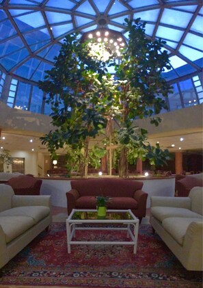 A spacious lobby with a high ceiling and a large tree in the center. There are sofas and a coffee table in the foreground. - Green Park Bologna Residence (Granarolo dell'Emilia)