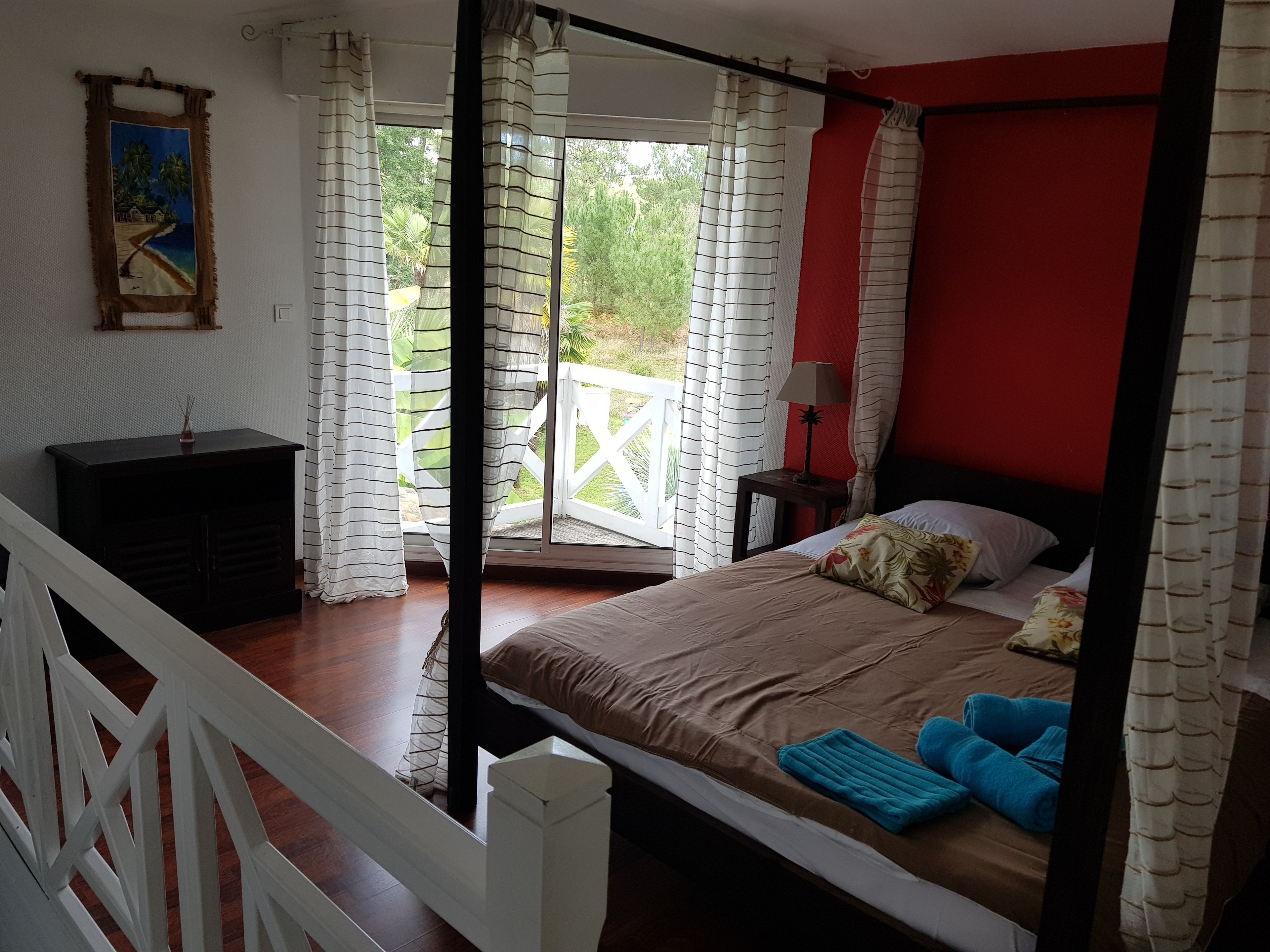 A bedroom with a bed, a nightstand, a lamp, a painting on the wall, and a view of greenery outside.