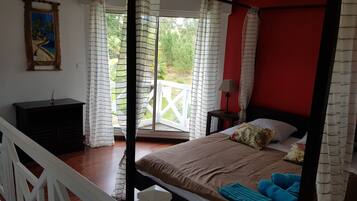 A bedroom with a bed, a nightstand, a lamp, a painting on the wall, and a view of greenery outside.