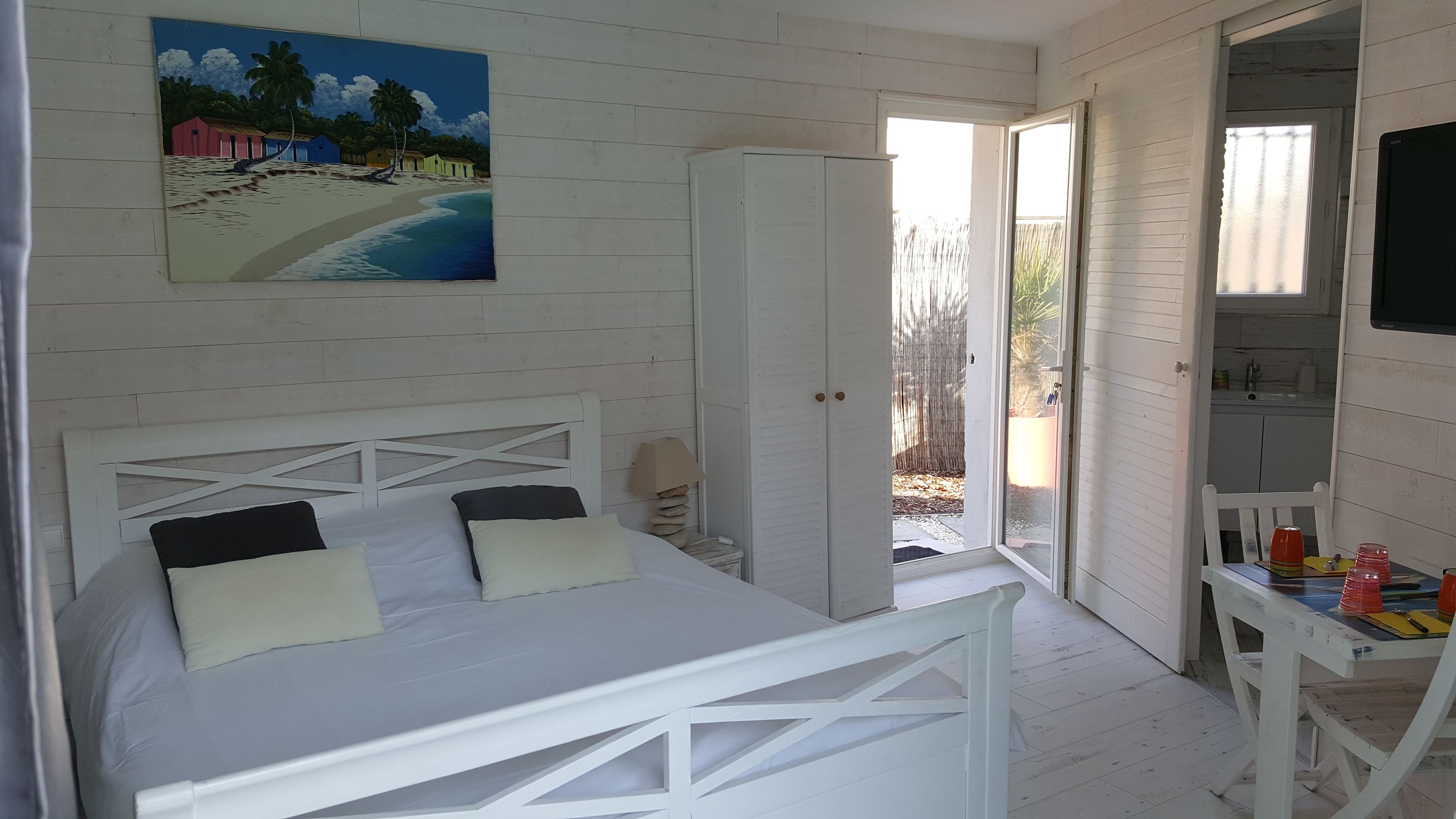 Et soveværelse med en hvid seng, et maleri med et strandmotiv og en dør der fører til en balkon.