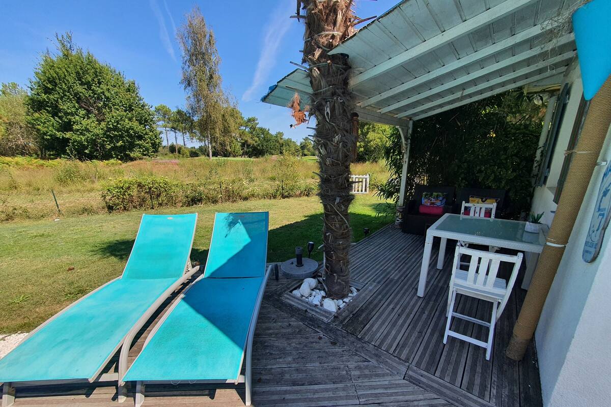 A wooden deck with two lounge chairs, a small table, and a covered area with a view of a grassy field and trees.
