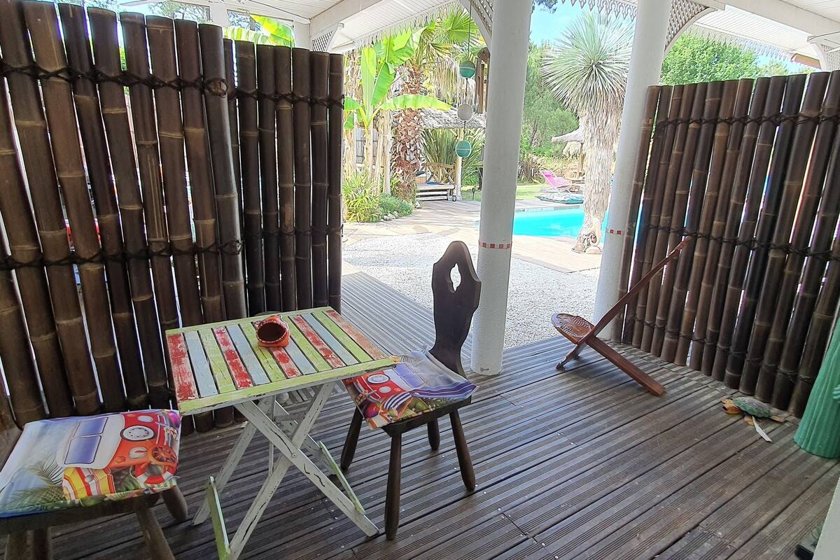 A wooden deck with a table and chairs, a bamboo fence, and a pool in the background.