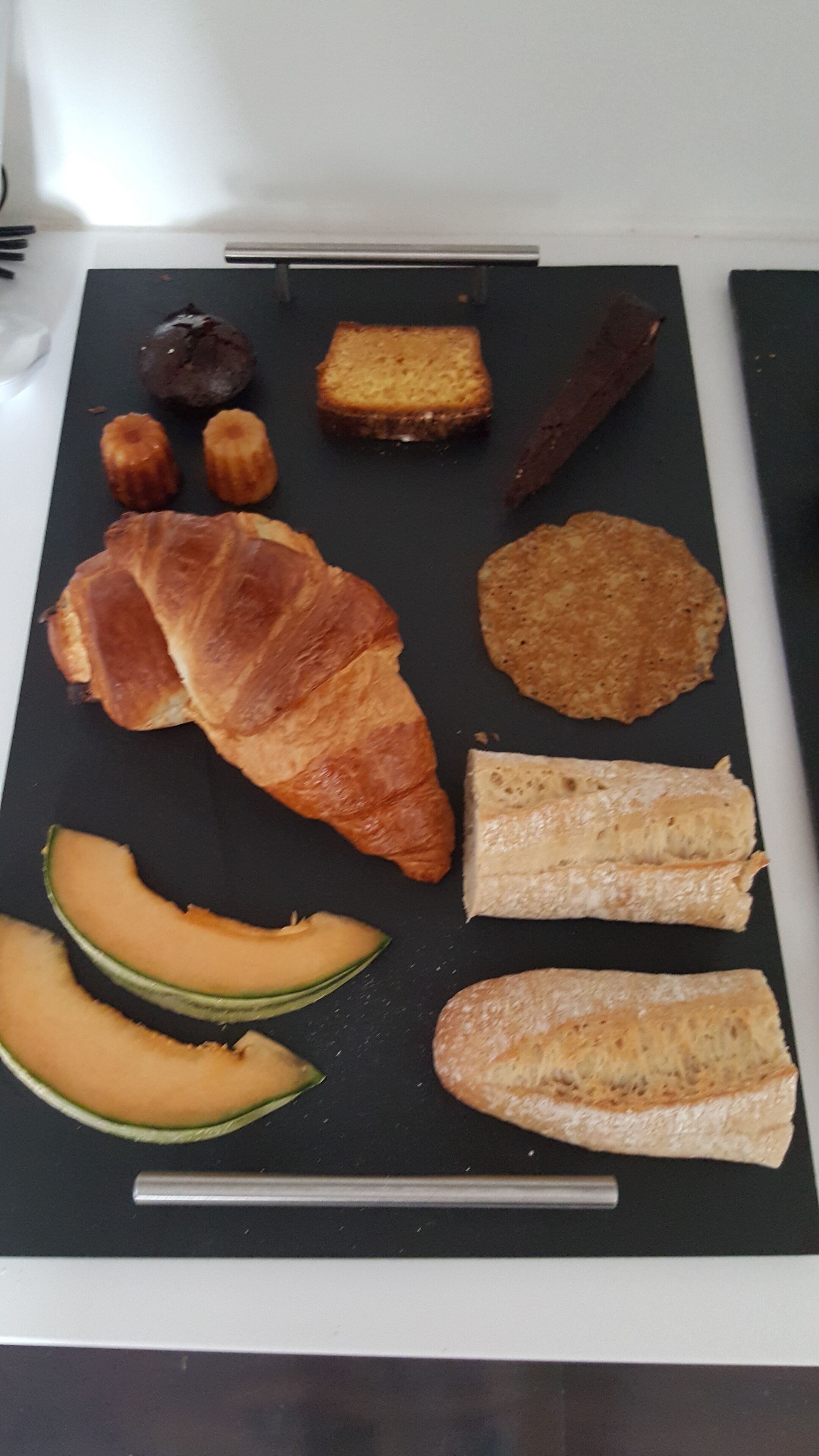 Verschiedene Speisen, darunter Croissants, Brot und Melonenscheiben, sind auf einer schwarzen Platte angerichtet.