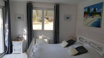 A bedroom with a bed, a window with curtains, a radiator, a small table with a lamp, and a painting on the wall.