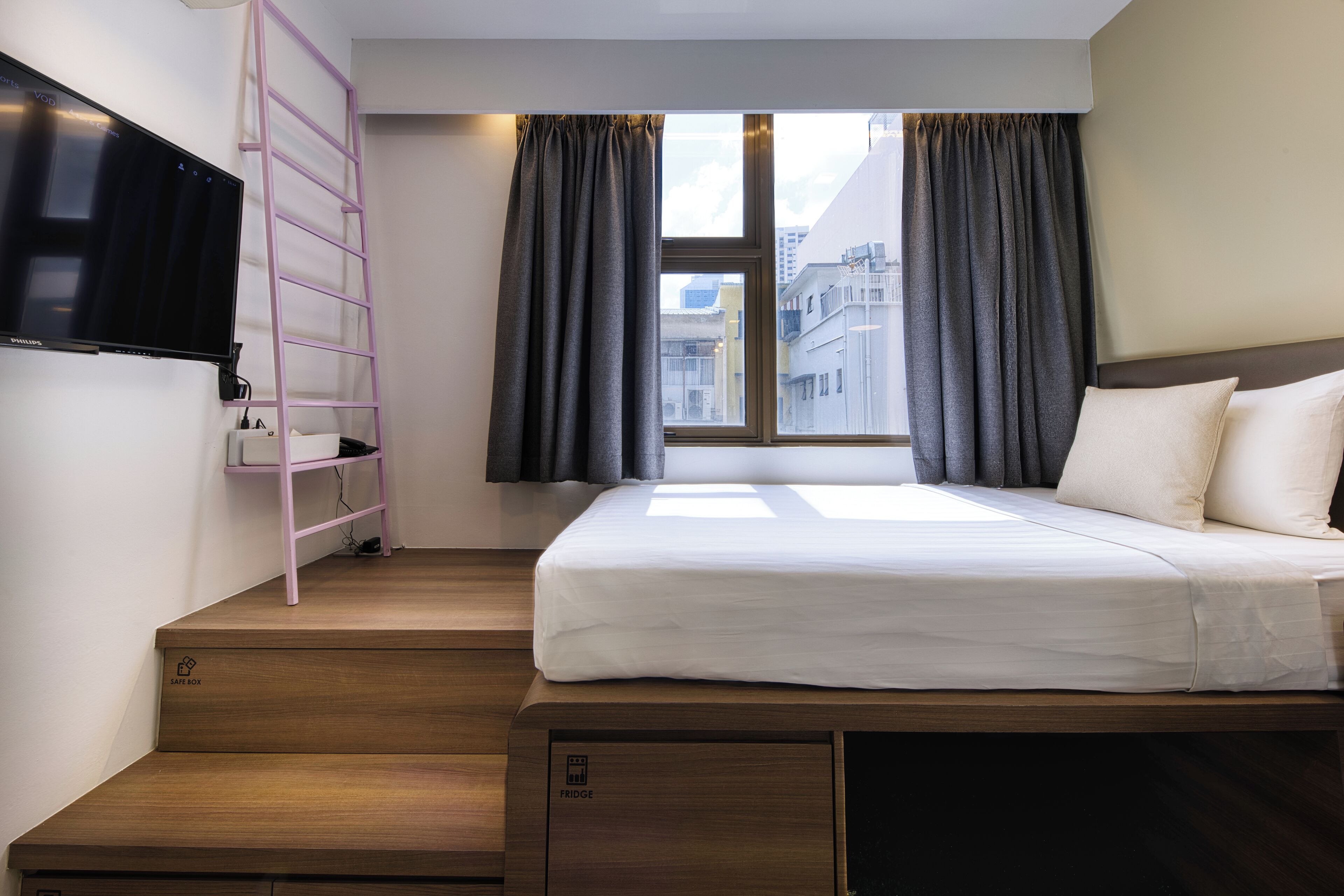 A hotel room with a bed, a TV mounted on the wall, a small shelf, a window with curtains, and a view of buildings outside.