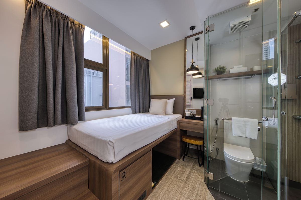 A compact hotel room with a bed, a desk, a chair, a shower, and a glass-enclosed bathroom.