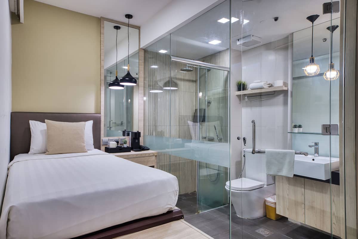A modern hotel room with a large bed, a glass-enclosed shower, a vanity with a mirror, and pendant lighting.