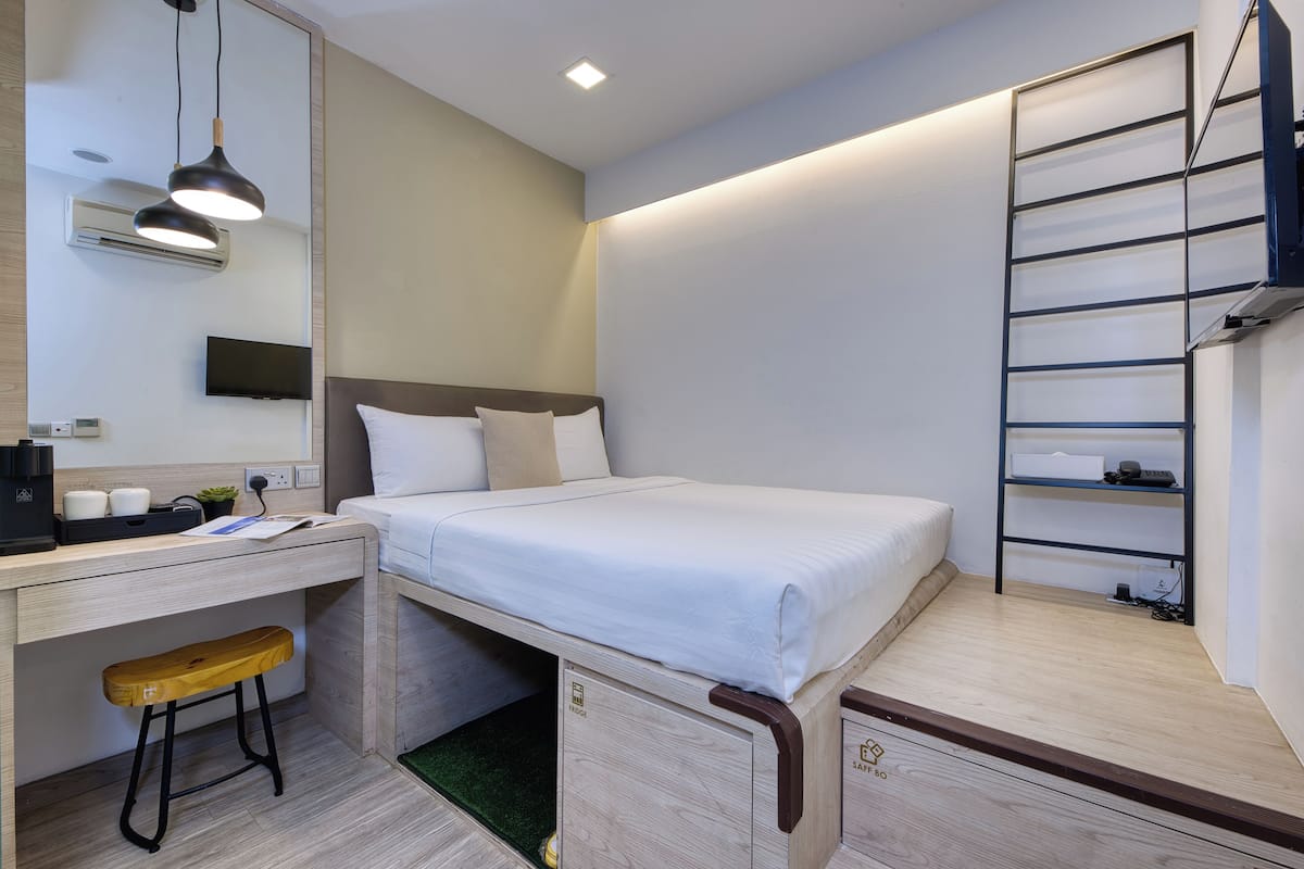 A compact hotel room with a bed, a desk, a chair, a TV, and a shelf.