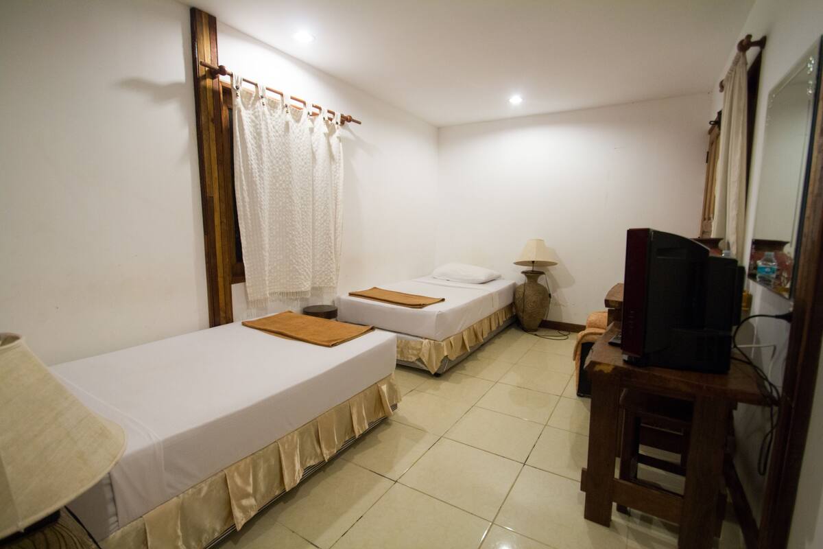 Une chambre d’hôtel avec deux lits simples, une petite table avec une lampe, un téléviseur posé sur un meuble en bois et un miroir au mur.