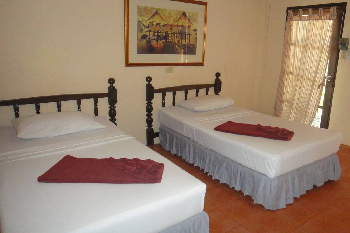 Une chambre avec deux lits, chacun doté d’un matelas blanc et d’une tête de lit en bois. Un tableau encadré est accroché au mur au-dessus des lits, et une fenêtre avec des rideaux se trouve à droite.