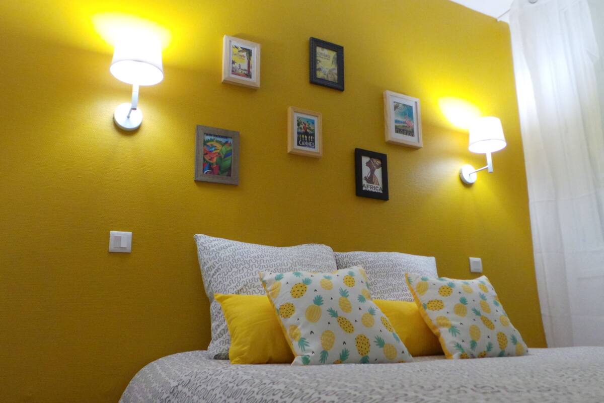 Una habitación con una pared amarilla, una cama con almohadas estampadas e iluminación montada en la pared.