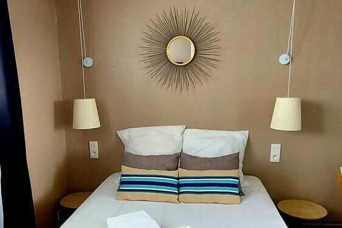 Una cama bien hecha con almohadas a rayas, dos lámparas de noche y un adorno de pared con diseño de sol.