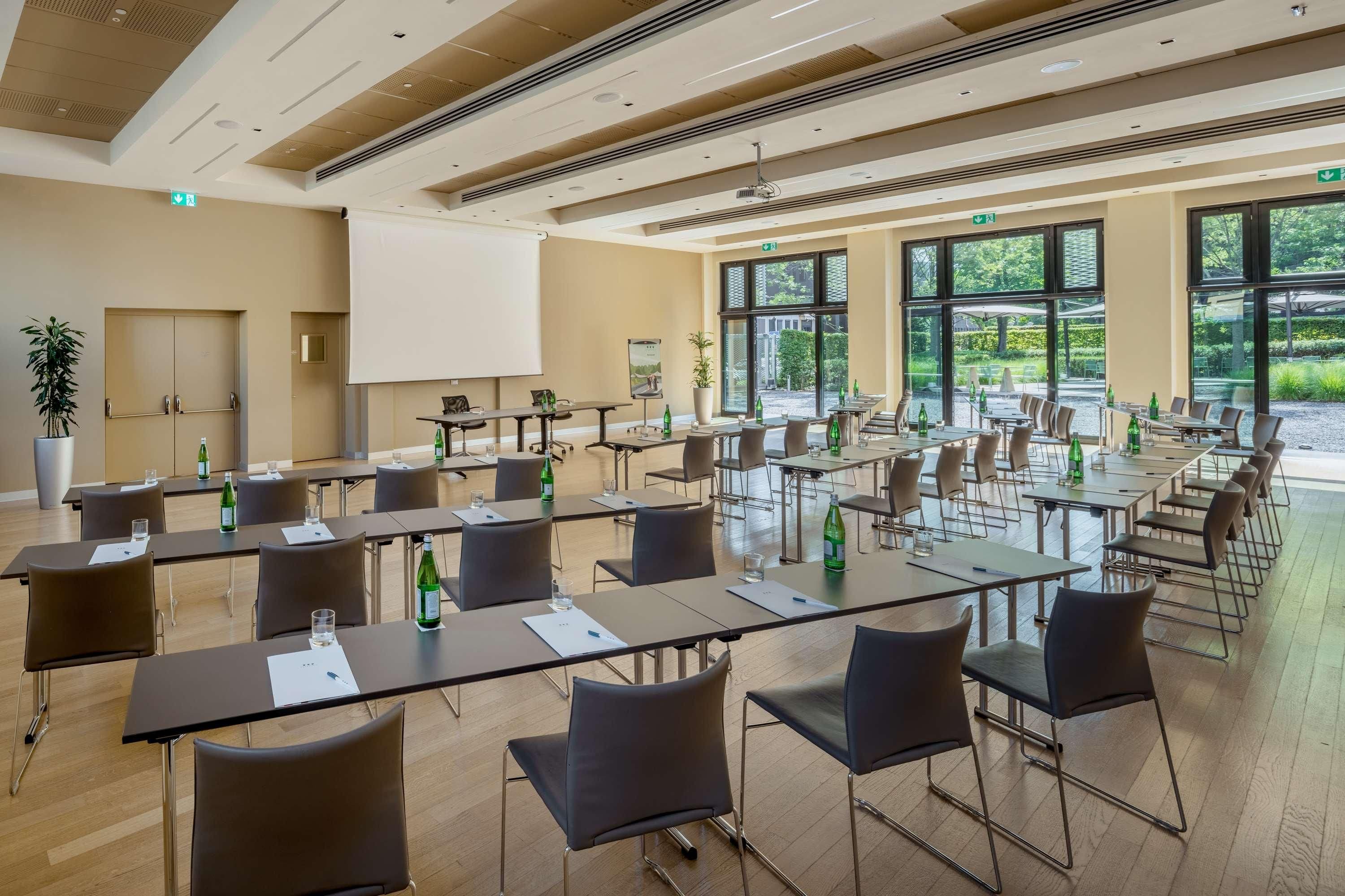 A conference room with multiple tables and chairs, a projector screen, and large windows with a view of the outdoors.