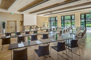 Un salón de conferencias con varias mesas y sillas, una pantalla de proyector y grandes ventanales con vistas al exterior.