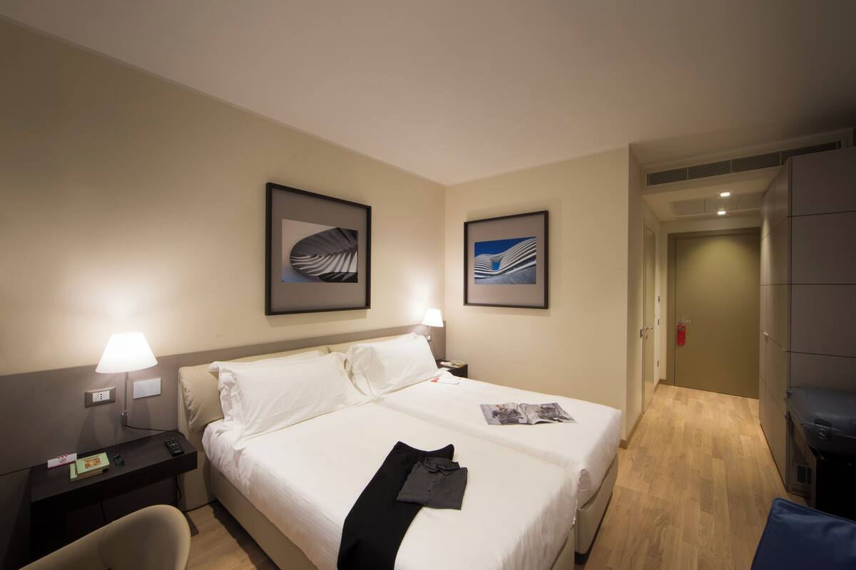 Hotellihuone, jossa on sänky, yöpöydät, työpöytä, tuoli ja kaksi kehystettyä kuvaa seinällä.