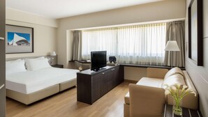 Una habitación de hotel moderna con una cama, un sofá, un televisor sobre un mueble de madera, un escritorio y un amplio ventanal con cortinas.
