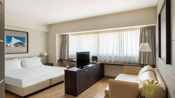 Ein modernes Hotelzimmer mit Bett, Sofa, Fernseher auf einer Holzkonsole, Schreibtisch und einem großen Fenster mit Vorhängen.